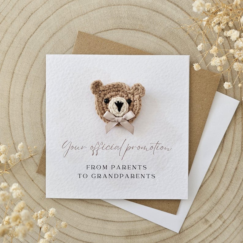 Puede incluir: Una tarjeta blanca con un oso de crochet marr&oacute;n con un lazo rosa. La tarjeta dice "Tu promoci&oacute;n oficial de padres a abuelos".