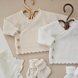 Puede incluir: Conjunto de ropa de beb&eacute; crema y blanca. El conjunto incluye dos c&aacute;rdigans cruzados de punto con botones de madera, una manta y dos pares de patucos. Los c&aacute;rdigans est&aacute;n en perchas decorativas. Los art&iacute;culos tienen un borde festoneado.