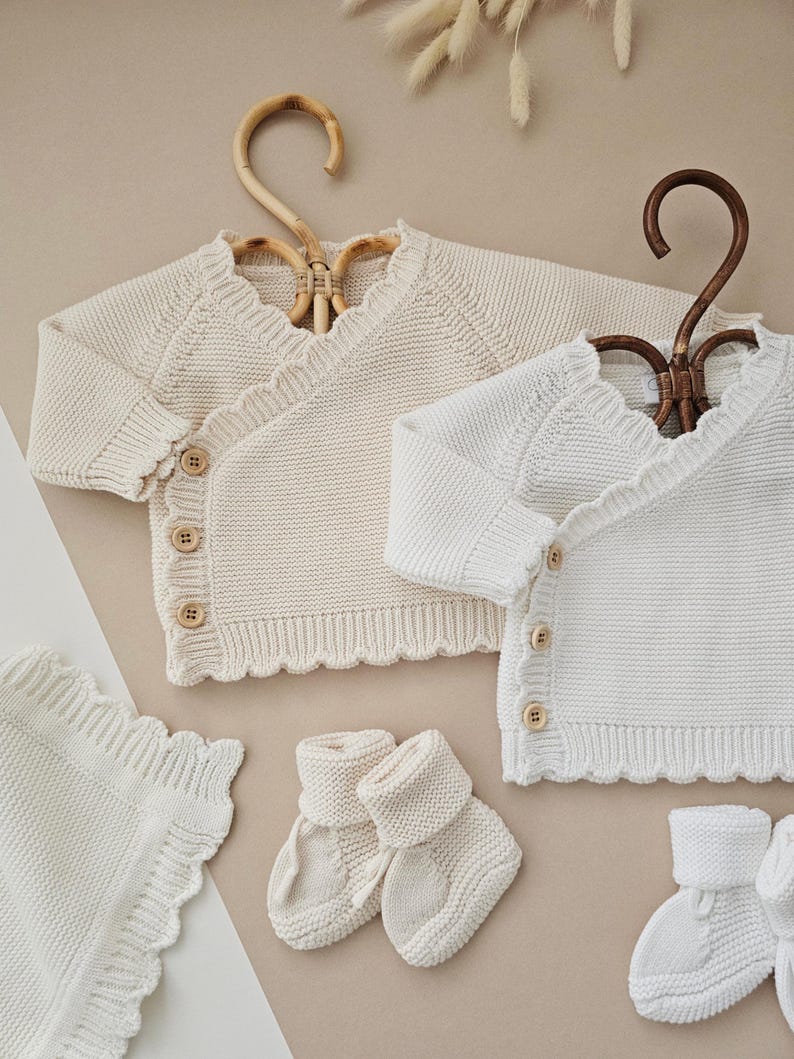 Puede incluir: Conjunto de ropa de beb&eacute; crema y blanca. El conjunto incluye dos c&aacute;rdigans cruzados de punto con botones de madera, una manta y dos pares de patucos. Los c&aacute;rdigans est&aacute;n en perchas decorativas. Los art&iacute;culos tienen un borde festoneado.