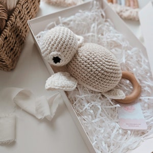 Könnte beinhalten: Ein gehäkeltes beigefarbenes Schildkrötenspielzeug mit einem Holzring. Das Spielzeug befindet sich in einer weißen Geschenkbox mit weißem Seidenpapier.