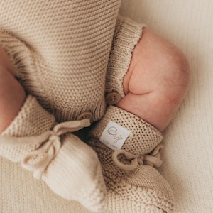 May include: A beige knitted romper with matching booties. The romper features a textured knit pattern and two small brown buttons at the leg opening. The booties have a tie closure and a white fabric label with the text "CY" and "WITH LOVE".