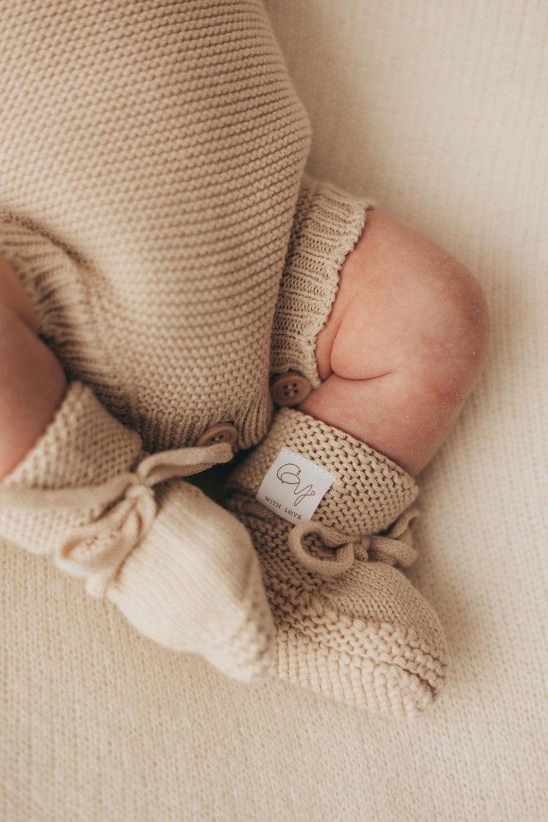 May include: A beige knitted romper with matching booties. The romper features a textured knit pattern and two small brown buttons at the leg opening. The booties have a tie closure and a white fabric label with the text "CY" and "WITH LOVE".