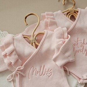 May include: Two light pink baby bodysuits with ruffled sleeves and wrap-around design, hanging on wooden hangers. One bodysuit has the name "Mollie" embroidered with a heart, and the other has the text "sweet she's lovely".