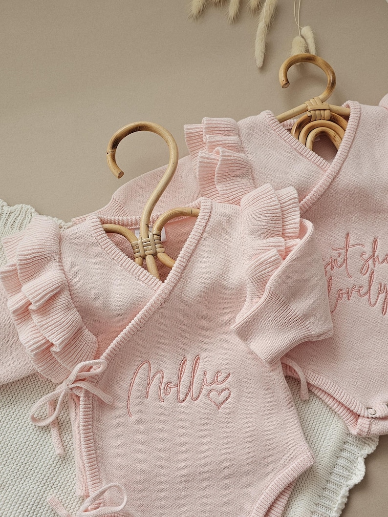 May include: Two light pink baby bodysuits with ruffled sleeves and wrap-around design, hanging on wooden hangers. One bodysuit has the name "Mollie" embroidered with a heart, and the other has the text "sweet she's lovely".