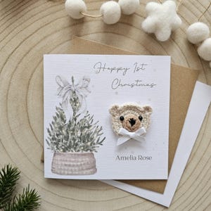 May include: A Christmas greeting card featuring a crocheted teddy bear and a festive tree illustration. The card says "Happy 1st Christmas" and "Amelia Rose". The card is on a wooden surface with felt decorations.