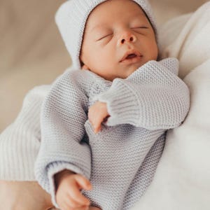 May include: A light grey knitted baby outfit consisting of a long-sleeved jumper with a textured garter stitch pattern and a matching beanie hat. The garment features ribbed cuffs and a ribbed brim on the hat, providing a soft and uniform appearance.