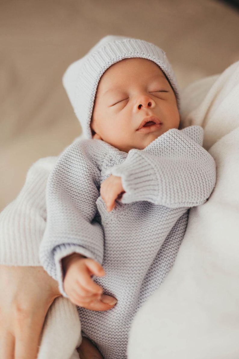 May include: A light grey knitted baby outfit consisting of a long-sleeved jumper with a textured garter stitch pattern and a matching beanie hat. The garment features ribbed cuffs and a ribbed brim on the hat, providing a soft and uniform appearance.