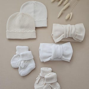Puede incluir: Un surtido de accesorios de beb&eacute; de punto blanco roto, que incluye dos gorros, dos diademas con detalles de lazo y dos pares de patucos con cierres de lazo. Los art&iacute;culos est&aacute;n dispuestos sobre un fondo neutro.