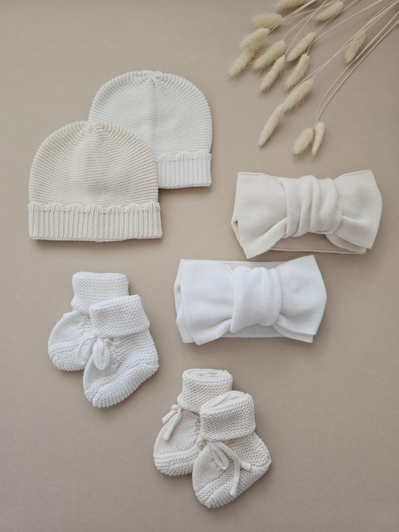 Puede incluir: Un surtido de accesorios de beb&eacute; de punto blanco roto, que incluye dos gorros, dos diademas con detalles de lazo y dos pares de patucos con cierres de lazo. Los art&iacute;culos est&aacute;n dispuestos sobre un fondo neutro.