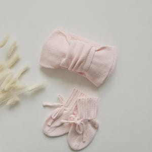 May include: A light pink baby headband with a large bow and a pair of matching baby booties with tie closures. The set is made of a soft, knitted material. The items are displayed on a white surface.