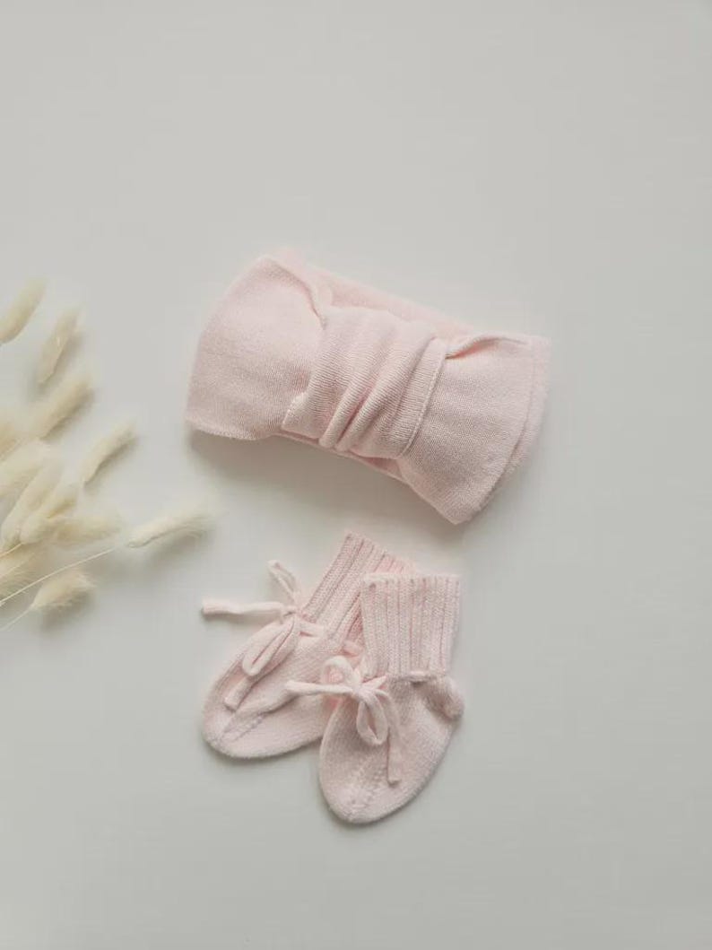 May include: A light pink baby headband with a large bow and a pair of matching baby booties with tie closures. The set is made of a soft, knitted material. The items are displayed on a white surface.