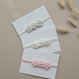 May include: Three baby headbands with crocheted flower details. The headbands are displayed on white cards. One is pale pink, one is white, and one is cream. Each flower has a pearl centre.