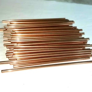 May include: A collection of numerous thin, cylindrical copper rods arranged in a horizontal stack. Each rod features a smooth, metallic, reddish-brown finish with a consistent diameter and straight edges, creating a uniform appearance across the group.