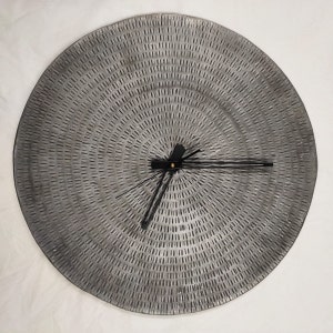 May include: A large, round, gray metal clock with a textured surface and black hands. The clock face has a concentric circle pattern.