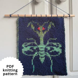 May include: A knitted wall hanging featuring a green praying mantis design on a dark blue background. The mantis has red eyes. The textile is suspended from a wooden dowel with a dark blue string. The text "PDF knitting pattern" is in the lower left corner.