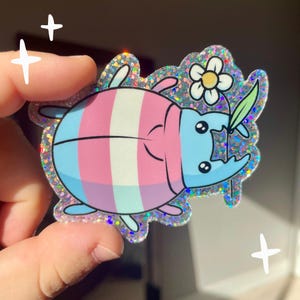 Puede incluir: Una pegatina brillante de un escarabajo de dibujos animados con una cáscara rayada de color rosa, azul y blanco. El escarabajo sostiene una flor blanca con un tallo verde.