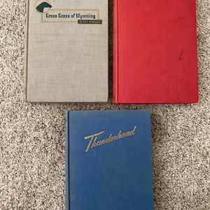 May include: Three hardcover books. The top left book is light grey with the title "Green Grass of Wyoming" and a horse graphic. The top right book is red. The bottom book is blue with the title "Thunderhead" in gold lettering.