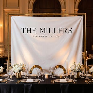 May include: Elegant wedding reception setup with a black tablecloth, gold accents, and white floral centerpieces. A white banner with "THE MILLERS" and the date "SEPTEMBER 20, 2026" is displayed. The scene includes candles, glassware, and gold chargers.
