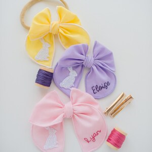 May include: Three fabric hair bows in yellow, lavender, and light pink. The yellow bow is on a beige headband. The lavender bow features an embroidered white rabbit and the name Eloise. The pink bow features an embroidered white rabbit and the name Ryan. Includes two spools of thread and two gold metal hair clips.