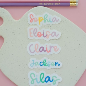 May include: Five die-cut name stickers featuring the names Sophia, Eloisa, Claire, Jackson, and Silas in colorful cursive fonts. Three pencils with the text THE BLOOMING BIKE and a heart symbol are positioned above the stickers on a speckled board.
