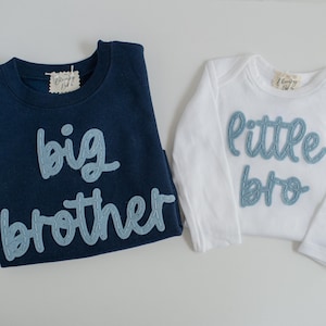 May include: Two children's shirts. One is navy blue with "big brother" in light blue script. The other is white with "little bro" in light blue script. Both shirts have a tag on the collar.
