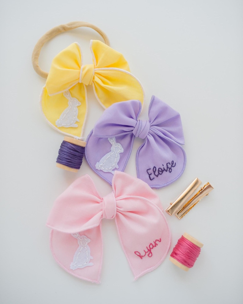 May include: Three fabric hair bows in yellow, lavender, and light pink. The yellow bow is on a beige headband. The lavender bow features an embroidered white rabbit and the name Eloise. The pink bow features an embroidered white rabbit and the name Ryan. Includes two spools of thread and two gold metal hair clips.