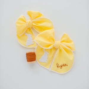May include: Two bright yellow fabric hair bows with white piped edges, each featuring a white embroidered rabbit silhouette. One bow includes the name Ryan stitched in brown thread. A small wooden spool of brown embroidery thread sits nearby.