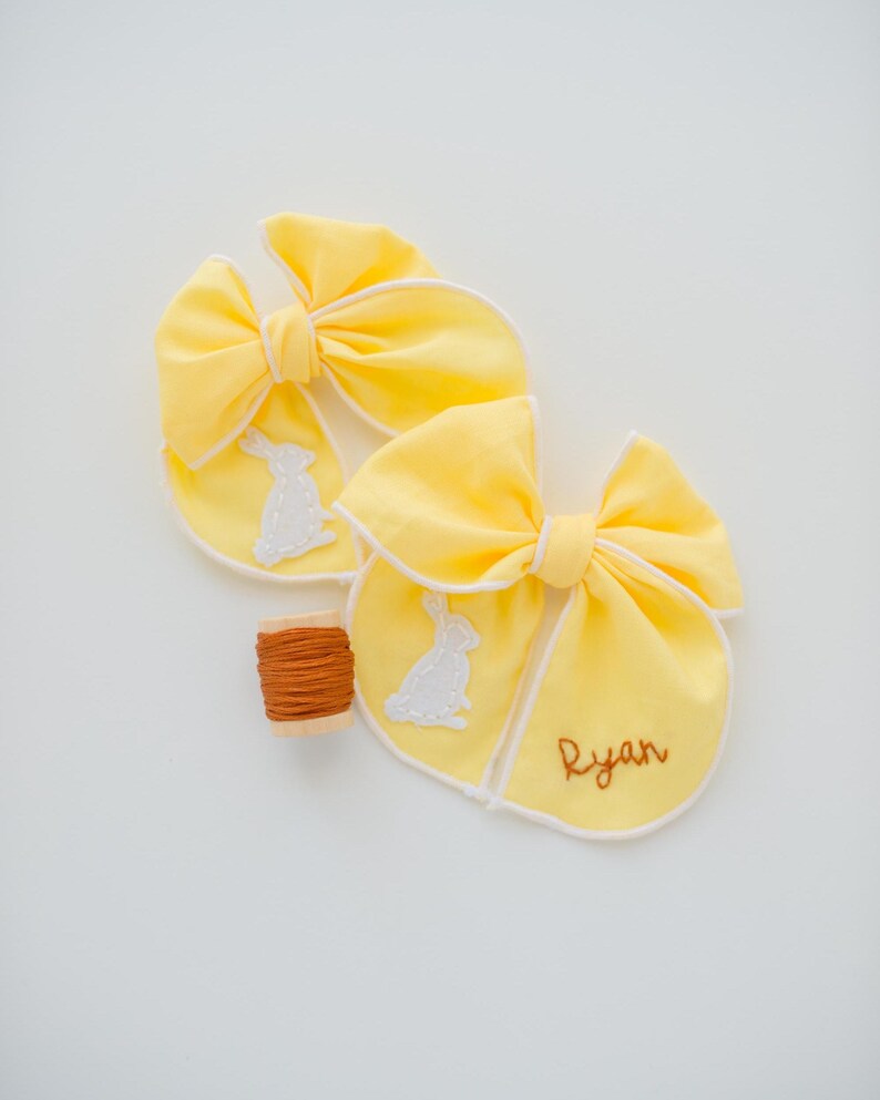 May include: Two bright yellow fabric hair bows with white piped edges, each featuring a white embroidered rabbit silhouette. One bow includes the name Ryan stitched in brown thread. A small wooden spool of brown embroidery thread sits nearby.