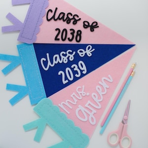 May include: Three triangular felt pennants in light pink, dark blue, and light pink. The top pennant features black text reading Class of 2038. The middle pennant has white text reading Class of 2039. The bottom pennant has white text reading mrs. green.