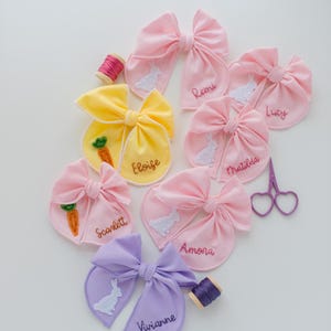 May include: A collection of fabric hair bows in pink, yellow, and lavender, each embroidered with a name and a white rabbit or orange carrot motif. Included are a small spool of pink thread, a spool of purple thread, and purple heart-shaped scissors.