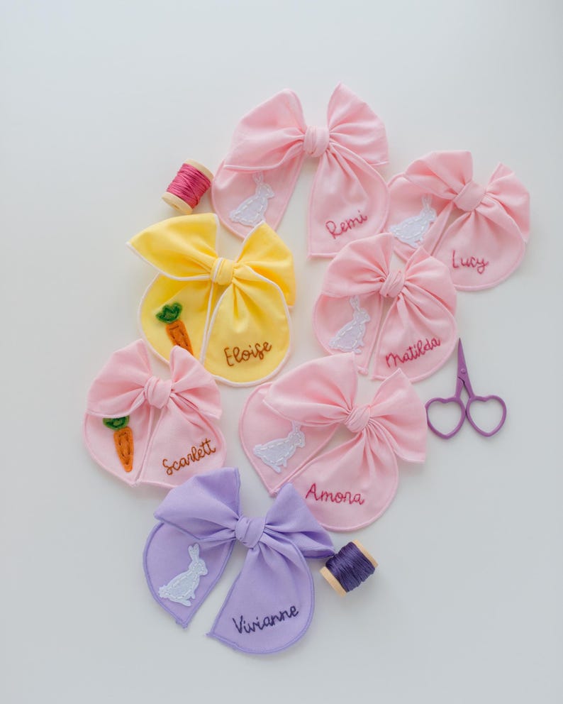 May include: A collection of fabric hair bows in pink, yellow, and lavender, each embroidered with a name and a white rabbit or orange carrot motif. Included are a small spool of pink thread, a spool of purple thread, and purple heart-shaped scissors.