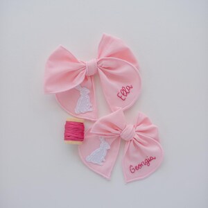 May include: Two light pink fabric hair bows featuring white stitched rabbit appliques and embroidered names Ella and Georgia in pink thread. A small wooden spool wrapped with matching pink embroidery floss is positioned between the two bows.
