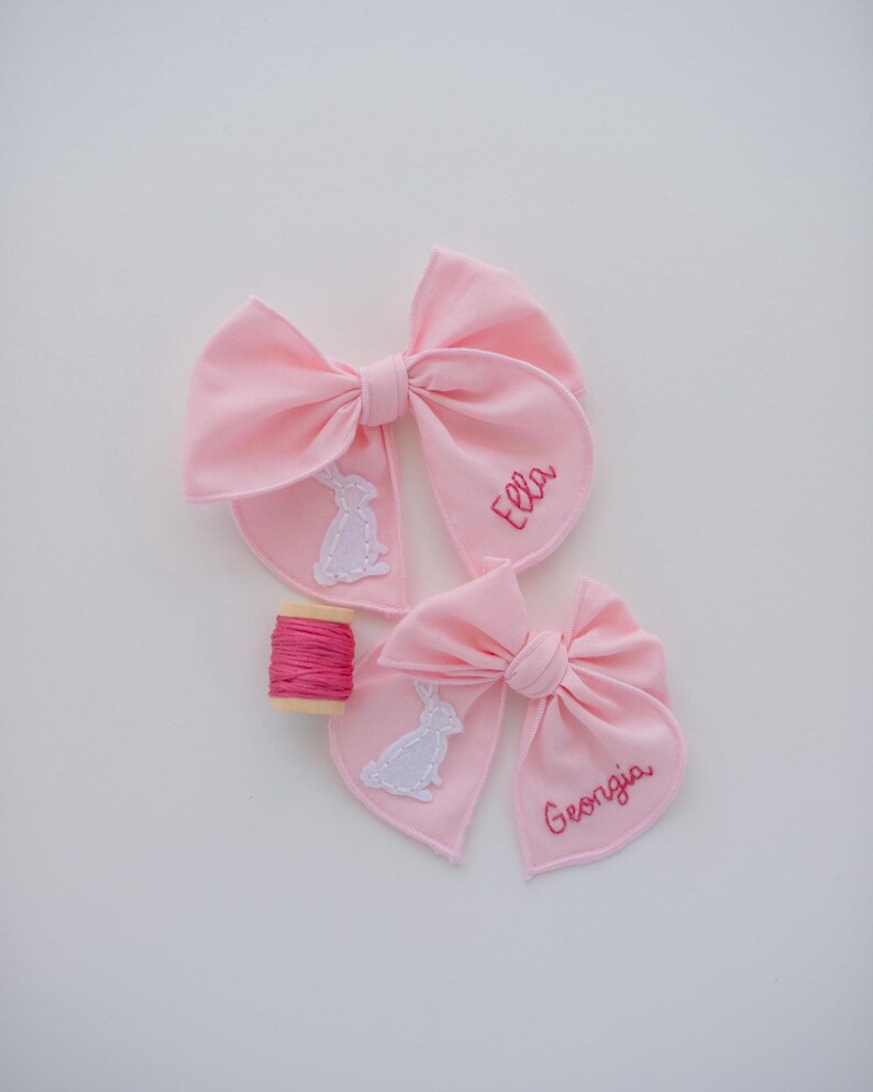 May include: Two light pink fabric hair bows featuring white stitched rabbit appliques and embroidered names Ella and Georgia in pink thread. A small wooden spool wrapped with matching pink embroidery floss is positioned between the two bows.