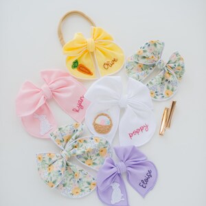 May include: A collection of fabric hair bows in yellow, pink, white, and lavender, featuring embroidered names like Olive, Ryan, Poppy, and Eloise. Some bows display stitched motifs of a carrot, a basket, and a rabbit, alongside floral patterns.