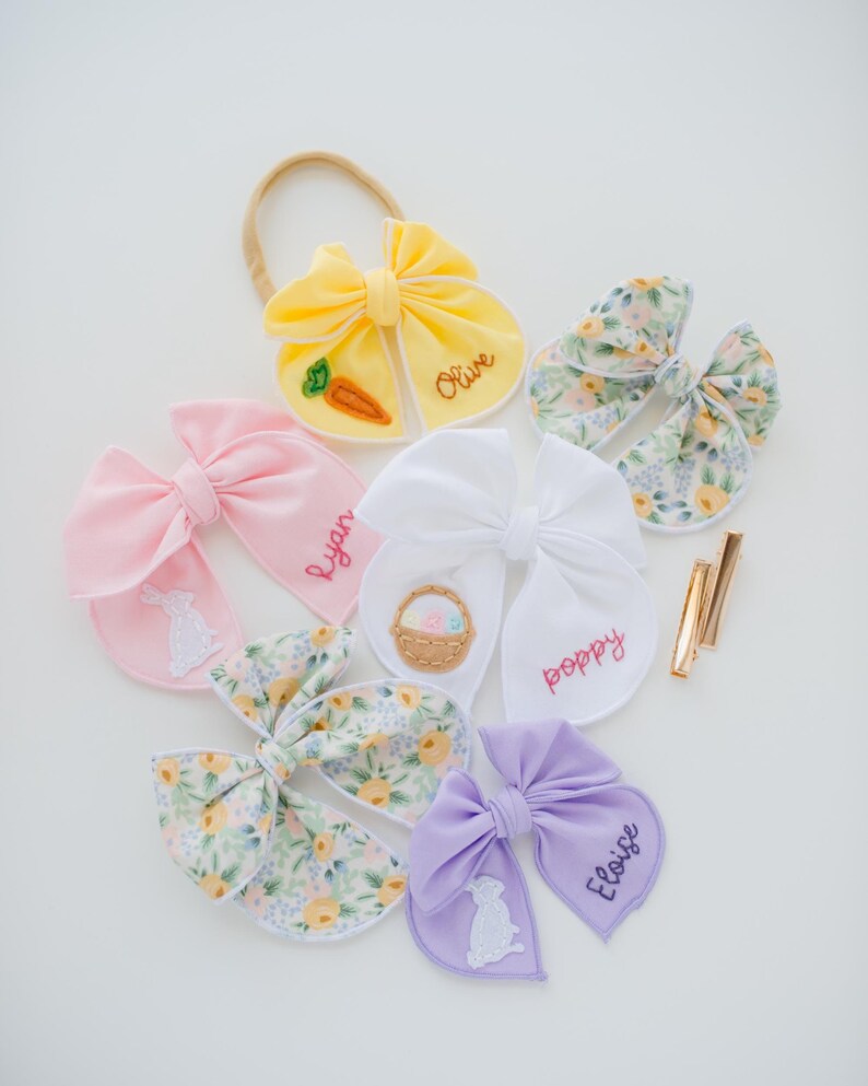 May include: A collection of fabric hair bows in yellow, pink, white, and lavender, featuring embroidered names like Olive, Ryan, Poppy, and Eloise. Some bows display stitched motifs of a carrot, a basket, and a rabbit, alongside floral patterns.
