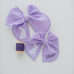May include: Two lavender fabric hair bows featuring white stitched rabbit silhouettes. One bow displays the name Eloise embroidered in dark purple thread. A small wooden spool wrapped with dark purple embroidery thread sits nearby.