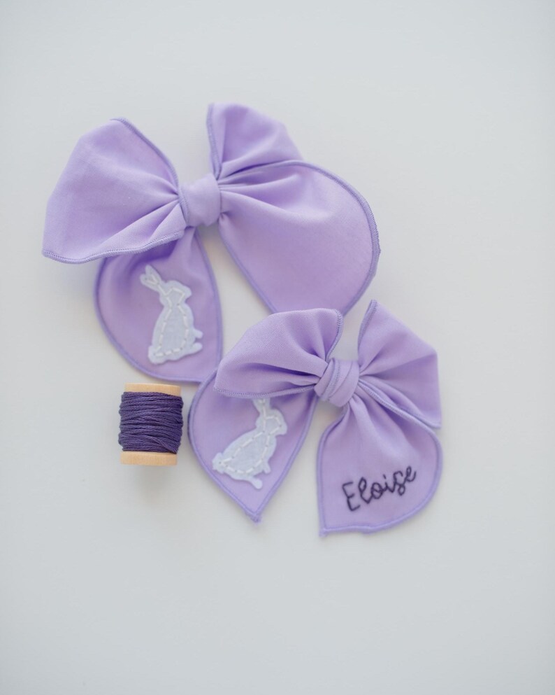 May include: Two lavender fabric hair bows featuring white stitched rabbit silhouettes. One bow displays the name Eloise embroidered in dark purple thread. A small wooden spool wrapped with dark purple embroidery thread sits nearby.