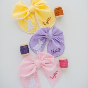 May include: Three fabric hair bows in yellow, lavender, and light pink, each featuring a white embroidered rabbit silhouette and a name stitched in cursive thread. The names Ryan and Eloise are visible, accompanied by three wooden spools of thread.