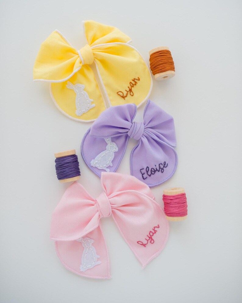 May include: Three fabric hair bows in yellow, lavender, and light pink, each featuring a white embroidered rabbit silhouette and a name stitched in cursive thread. The names Ryan and Eloise are visible, accompanied by three wooden spools of thread.