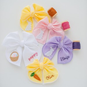 May include: A collection of five fabric hair bows in yellow, pink, white, lavender, and light yellow. The bows feature embroidered bunny, carrot, and Easter basket motifs, along with the names Poppy, Eloise, and Olive stitched in thread.