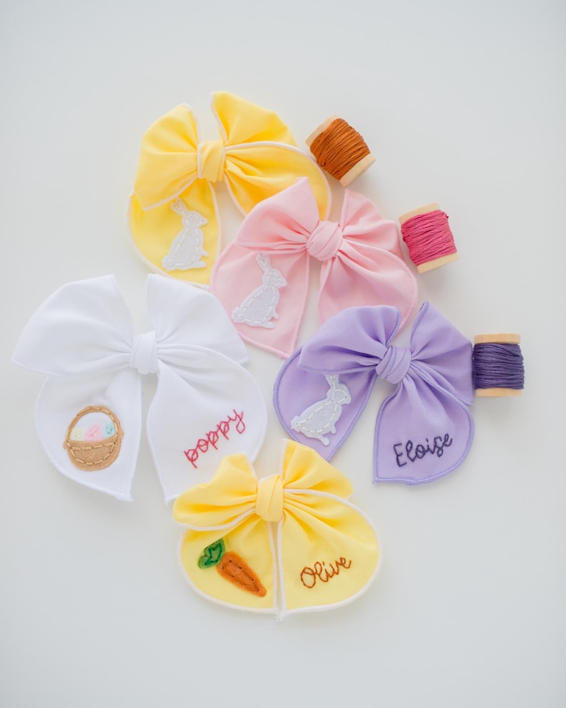 May include: A collection of five fabric hair bows in yellow, pink, white, lavender, and light yellow. The bows feature embroidered bunny, carrot, and Easter basket motifs, along with the names Poppy, Eloise, and Olive stitched in thread.