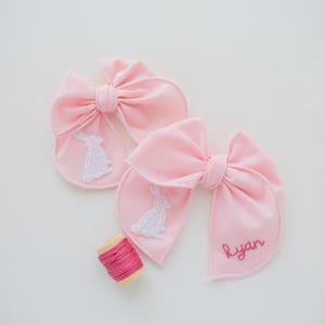 May include: Two light pink fabric bows featuring white embroidered rabbit silhouettes on the tails. One bow includes the name Ryan embroidered in dark pink thread. A small wooden spool of matching dark pink embroidery thread sits nearby.