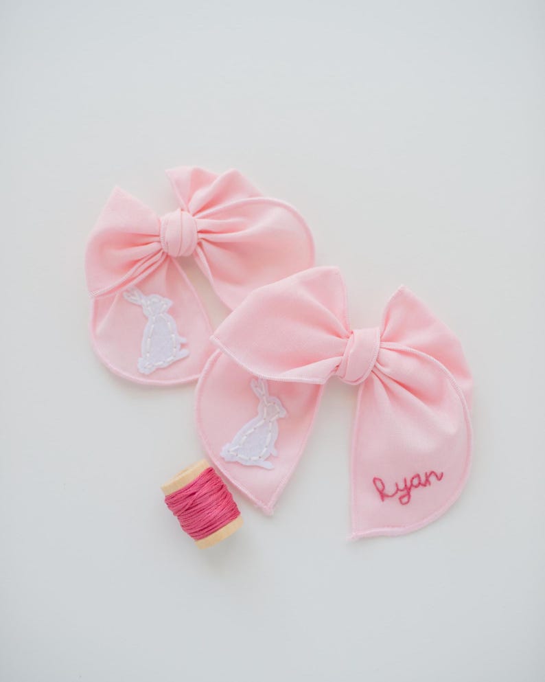 May include: Two light pink fabric bows featuring white embroidered rabbit silhouettes on the tails. One bow includes the name Ryan embroidered in dark pink thread. A small wooden spool of matching dark pink embroidery thread sits nearby.