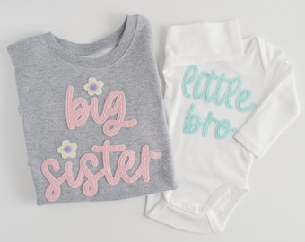 May include: A gray long-sleeve shirt with the words "big sister" in pink, and a white onesie with "little bro" in turquoise. Both have decorative stitching and flower accents, perfect for siblings.