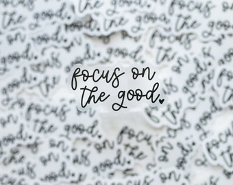 May include: A clear sticker with the text "focus on the good" in black cursive lettering with a small heart symbol. The sticker is surrounded by many other stickers with the same text in the background.