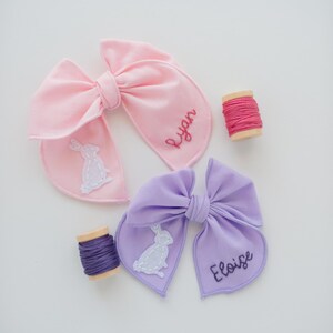 May include: A light pink hair bow embroidered with the name Ryan and a white rabbit, and a light purple hair bow embroidered with the name Eloise and a white rabbit, accompanied by two small wooden spools of matching pink and purple embroidery thread.