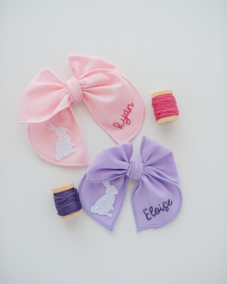 May include: A light pink hair bow embroidered with the name Ryan and a white rabbit, and a light purple hair bow embroidered with the name Eloise and a white rabbit, accompanied by two small wooden spools of matching pink and purple embroidery thread.