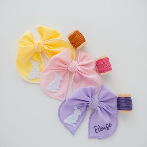 May include: Three fabric hair bows in yellow, pink, and lavender, each featuring a white embroidered rabbit silhouette. The lavender bow includes the name Eloise embroidered in dark thread. Three wooden spools of matching embroidery thread are included.