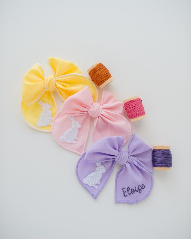 May include: Three fabric hair bows in yellow, pink, and lavender, each featuring a white embroidered rabbit silhouette. The lavender bow includes the name Eloise embroidered in dark thread. Three wooden spools of matching embroidery thread are included.