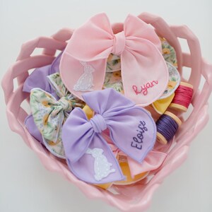 May include: A pink heart-shaped basket containing fabric bows in pink, lavender, and floral patterns. The bows feature embroidered white rabbit silhouettes and the names Ryan and Eloise stitched in thread. Two small wooden spools of thread are included.